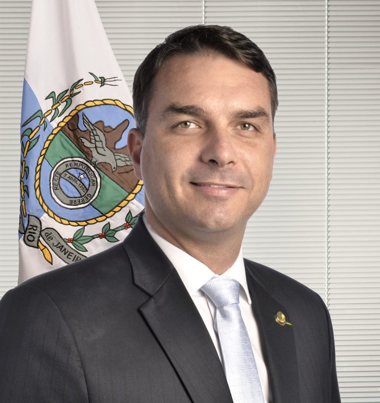 Flávio Bolsonaro em retrato oficial, de terno, com bandeira do Rio de Janeiro ao fundo. Foto: Agência Senado, 2019